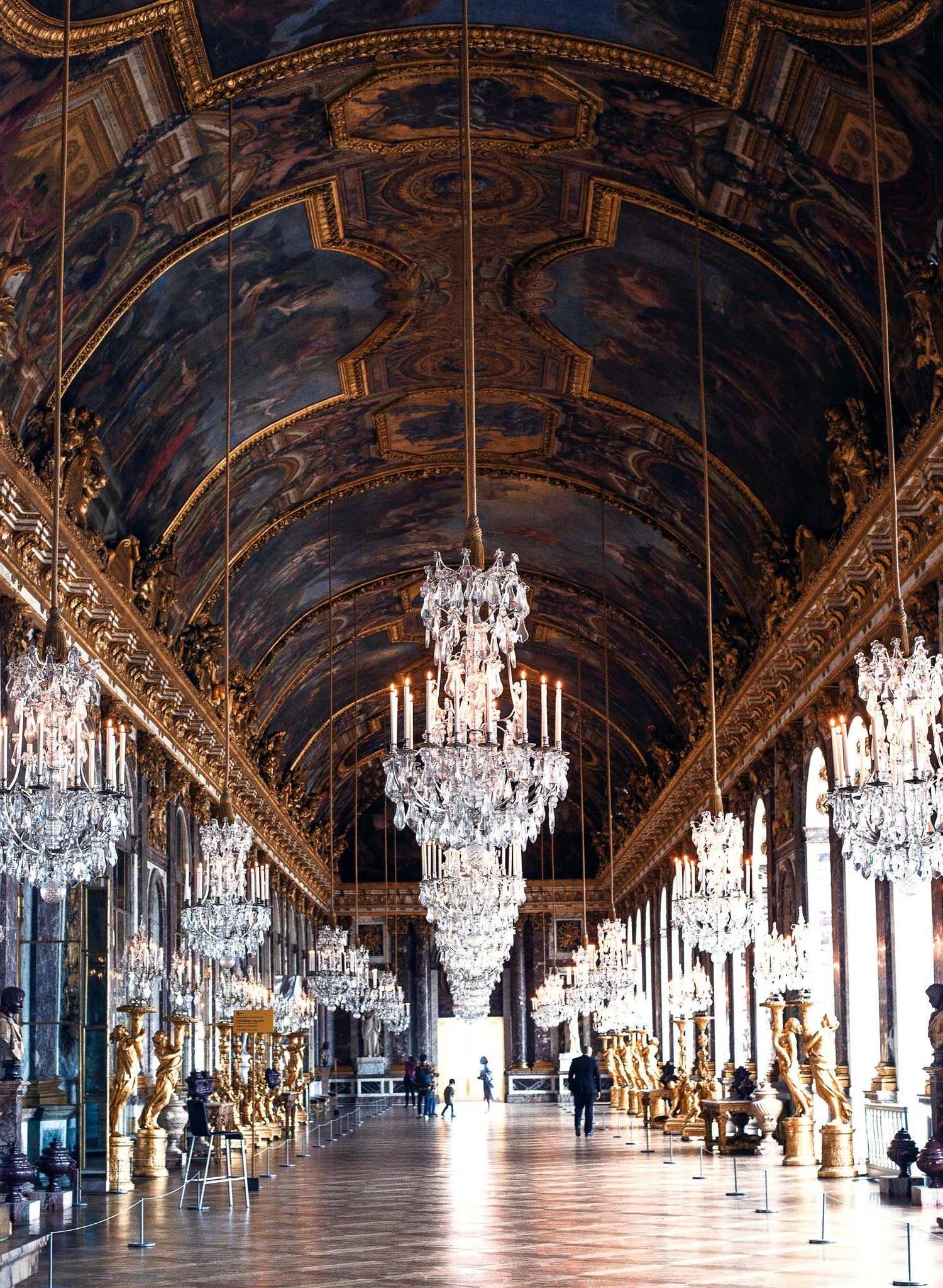 Grand hall with ornate gold sculptures, large crystal chandeliers, arched windows, and a painted vaulted ceiling